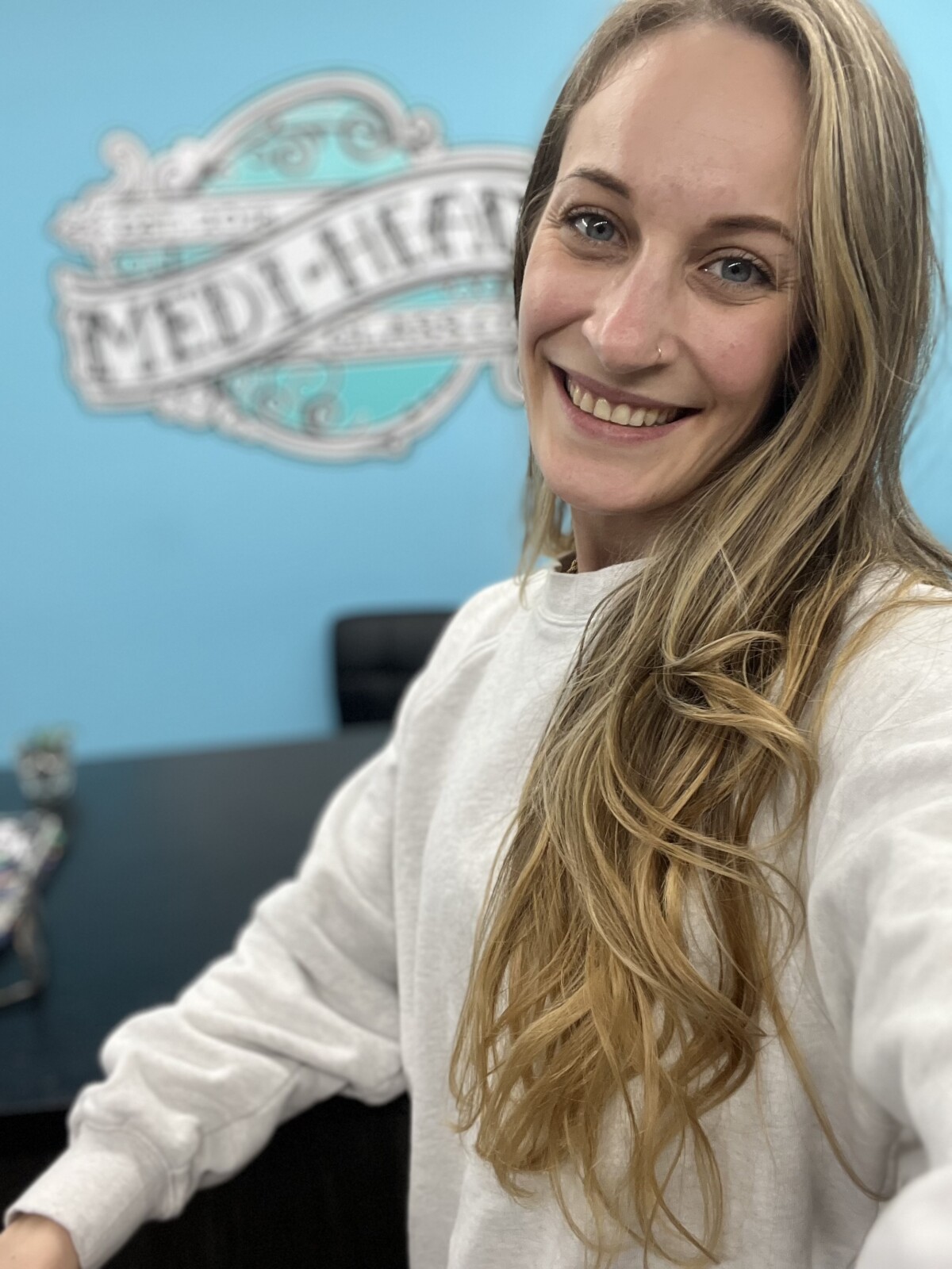 Kailee Hardie, Owner of Medi Heady Glass Co., wearing a white sweatshirt, takes a selfie in an office with a blue wall featuring the logo of Medi Heady Glass Co., which specializes in creating heady glass for cannabis concentrates.